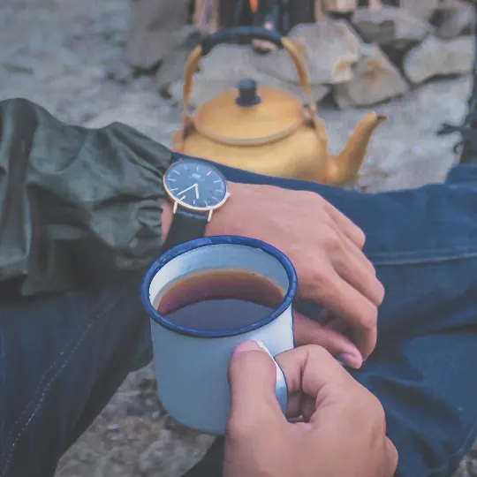 Someone sitting on a rock with an enamel mug of coffee, a kettle behind
